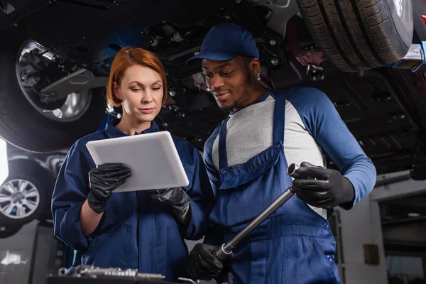 Multiethnic mechanics in gloves using digital tablet and holding wrench in car service — Stockfoto