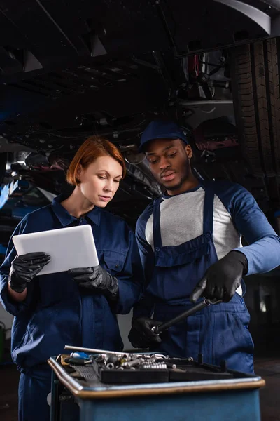 Mecánico afroamericano apuntando a herramientas cercanas a colega con tableta digital en servicio de automóvil - foto de stock