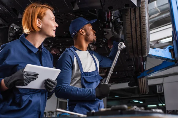 Side view of smiling multiethnic mechanics working with wrench, digital tablet and car in garage — Stock Photo