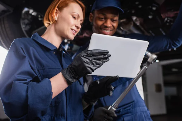 Tableta digital en la mano del mecánico cerca borroso africano americano colega con llave inglesa en servicio de coche - foto de stock