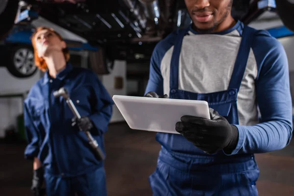 Afrikanischer Mechaniker mit digitalem Tablet in der Nähe von verschwommenem Kollegen und Auto in Garage — Stockfoto