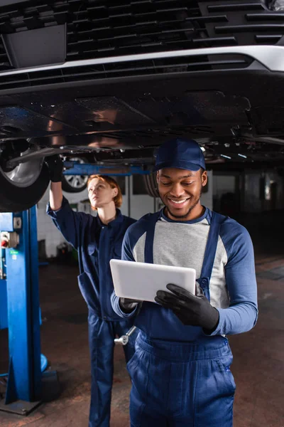 Lächelnder afrikanischer Mechaniker in Handschuhen mit digitalem Tablet im Autoservice — Stockfoto