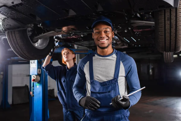 Mecânico afro-americano sorridente com tablet digital olhando para a câmera perto de colega e carro na garagem — Fotografia de Stock