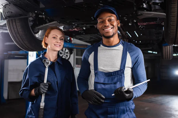 Positive multiethnic mechanics holding wrench and digital tablet near car in garage — Fotografia de Stock