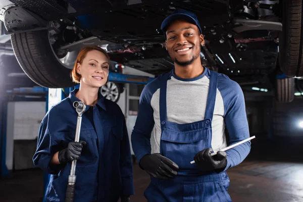 Positive multiethnic mechanics holding wrench and digital tablet near car in garage — Stock Photo
