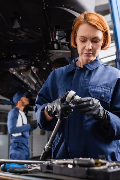 Mecánico en guantes que sostiene la llave inglesa cerca de herramientas borrosas en el servicio de automóviles - foto de stock