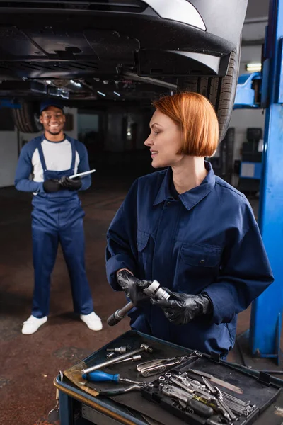 Delantera sonriente con guantes sosteniendo una llave inglesa cerca de un borroso colega afroamericano y un coche en el garaje - foto de stock