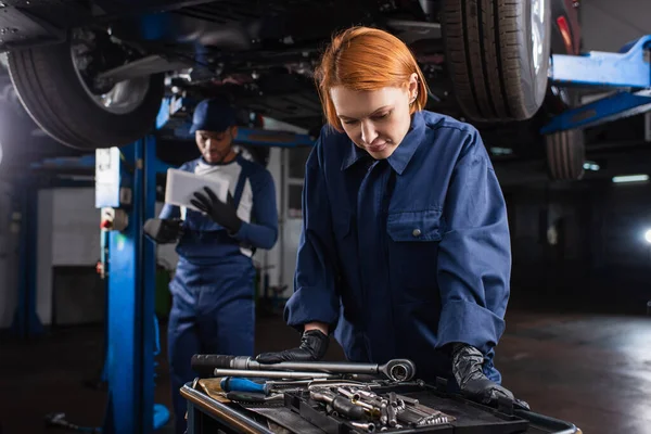 Mechaniker in Handschuhen bei der Suche nach Werkzeugen in der Nähe verschwommener afrikanischer amerikanischer Kollege und Dienstwagen — Stockfoto