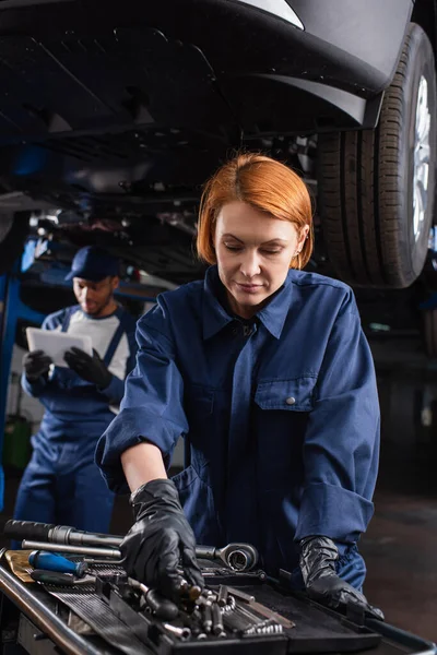 Mechanic in uniform taking tool near blurred african american colleague with digital tablet and car in garage — Stock Photo