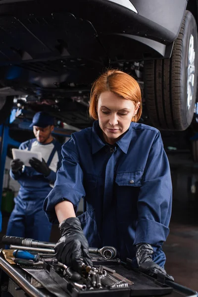 Mechanic in uniform taking tool near blurred african american colleague with digital tablet and car in garage — Foto stock