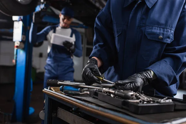 Mechanic in gloves taking tool near blurred african american colleague in garage — Stock Photo