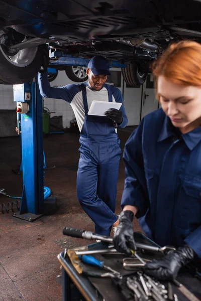 Mecánico afroamericano mirando tableta digital cerca de coche y colega borroso en servicio - foto de stock