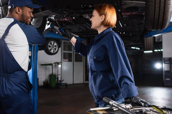 Vista laterale di allegra meccanica multietnica che lavora con auto vicino a utensili in garage — Foto stock