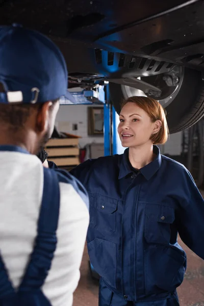 Lächelnder Mechaniker in Uniform im Gespräch mit verschwommenem afrikanisch-amerikanischem Kollegen unter Auto in Garage — Stockfoto