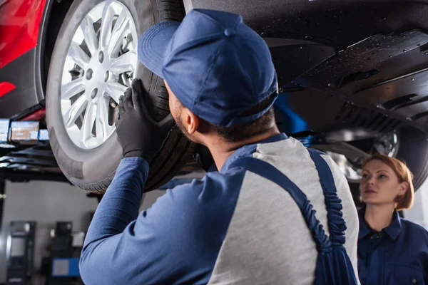 African american mechanic looking at car wheel near colleague in garage — стоковое фото