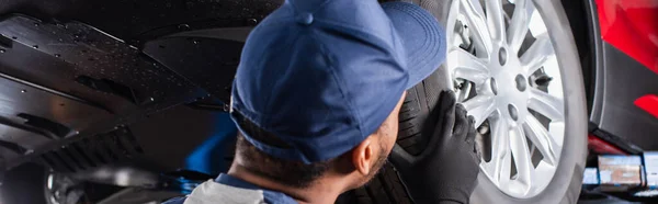 African american mechanic in cap looking at car wheel in garage, banner — Foto stock