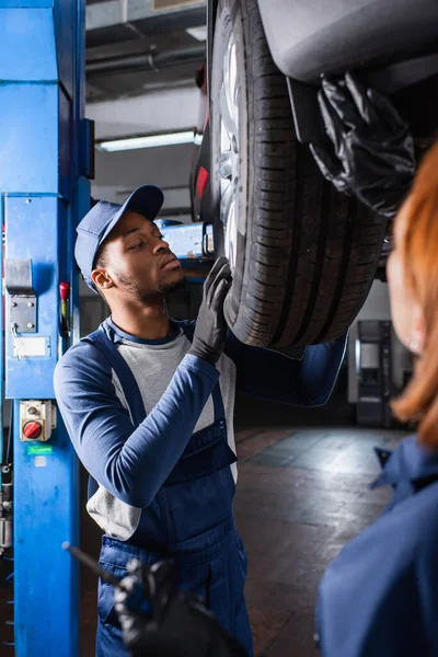 Afro-americano reparador trabajando con rueda de coche cerca borrosa colega en servicio - foto de stock