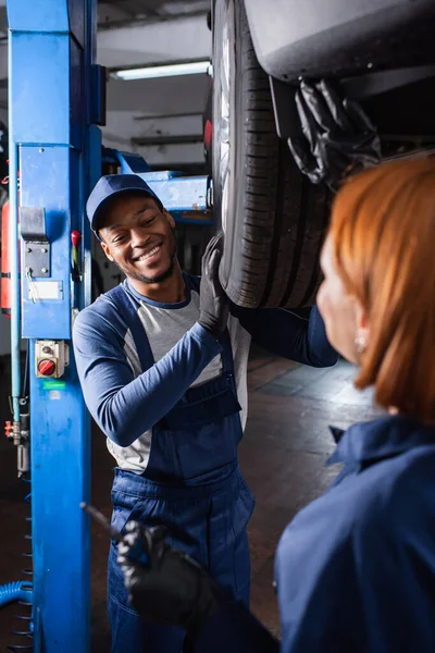 Positive african american mechanic working with car wheel and blurred repairwoman in service — Fotografia de Stock