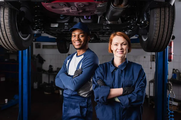 Sonriente mecánica interracial mirando a la cámara cerca del coche en el garaje - foto de stock