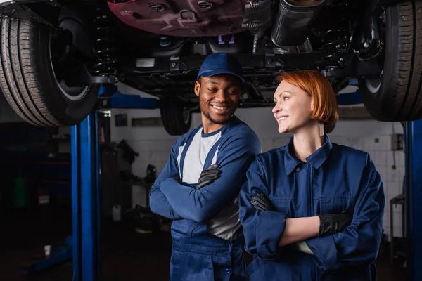 Mecánica multiétnica sonriente cruzando brazos cerca de coche en servicio - foto de stock