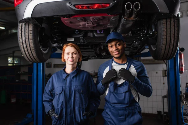 Multiethnic mechanics in uniform looking at camera under car in service — Stockfoto