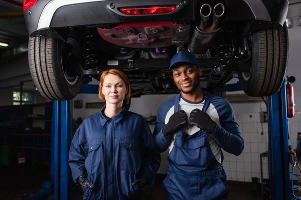 Multiethnic mechanics in uniform looking at camera under car in service — Stock Photo