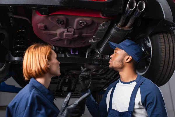 Vista de ángulo bajo de la mecánica multiétnica hablando bajo el coche en el garaje - foto de stock