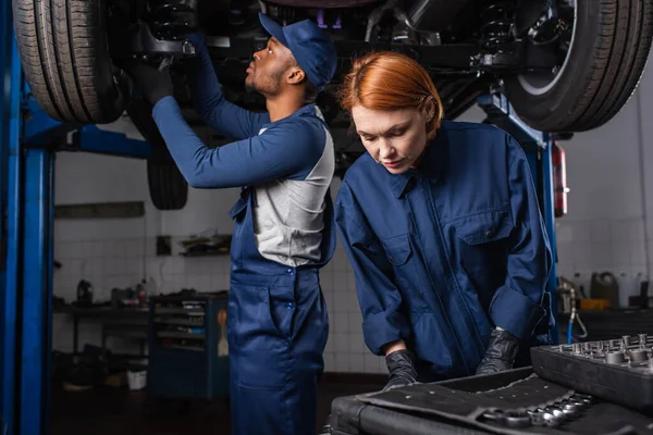 Механік стоїть біля інструментів і афроамериканський колега, який працює з машиною в гаражі — стокове фото