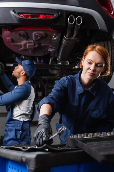 Réparatrice prenant clé tandis que le collègue afro-américain travaillant avec l'automobile dans le garage — Photo de stock