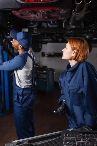 Seitenansicht eines rothaarigen Mechanikers, der einen Schraubenschlüssel in der Nähe eines afrikanisch-amerikanischen Kollegen hält, der mit einem Dienstwagen arbeitet — Stockfoto