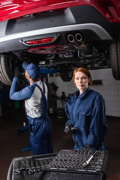 Redhead mechanic holding tool and looking at camera near car and african american colleague in garage — Stockfoto