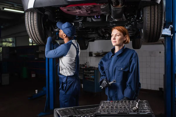 Mechanic in uniform holding wrench while african american colleague working with car in garage — Stock Photo