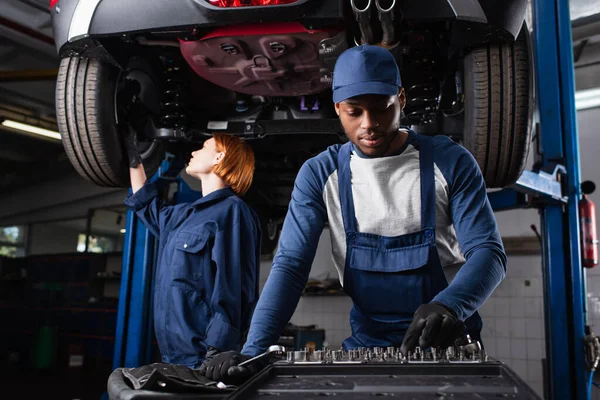 Africano americano mecánico tomando herramienta cerca colega trabajando con coche en garaje - foto de stock