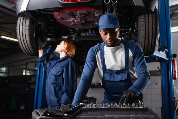 Meccanico afroamericano che prende utensile vicino collega che lavora con auto in garage — Foto stock