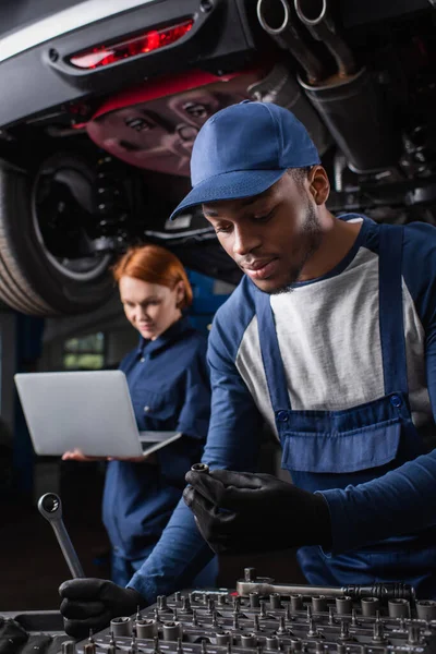 Africano americano mecánico herramientas de retención mientras que el colega que utiliza el ordenador portátil cerca de coche en el garaje - foto de stock