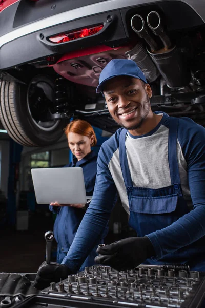 Smiling african american mechanic holding tools near colleague using laptop in car service — Stock Photo