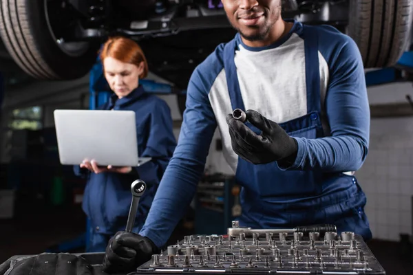 Africano americano reparador segurando ferramentas perto colega borrado com laptop no serviço de carro — Fotografia de Stock