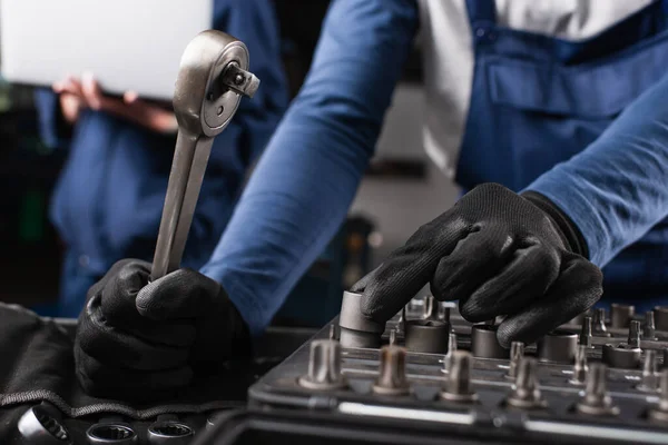 Ausgeschnittene Ansicht eines Mechanikers, der Schraubenschlüssel in der Nähe verschwommener Kollegen mit Laptop in der Garage hält — Stockfoto