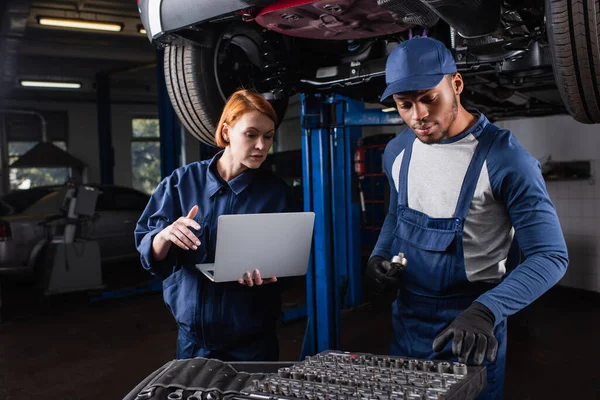 Mechanic holding laptop and talking to african american colleague with tools in car service — Fotografia de Stock