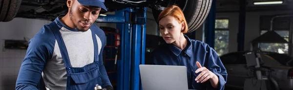 Workwoman holding laptop near african american colleague with wrench in car service, banner — Stock Photo