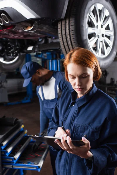 Mechanische Schrift auf Klemmbrett in der Nähe verschwommener afrikanischer amerikanischer Kollege und Auto in Garage — Stockfoto