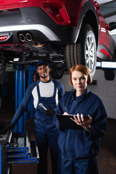 Multiethnic mechanics with clipboard and tools looking at camera near car in garage — Foto stock