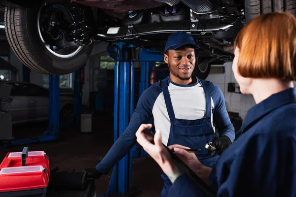 Lächelnder afrikanisch-amerikanischer Mechaniker mit Schraubenschlüssel im Gespräch mit einem Kollegen mit Klemmbrett im Autoservice — Stockfoto