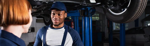Lächelnder afrikanisch-amerikanischer Mechaniker blickt verschwommenen Kollegen im Autoservice an, Banner — Stockfoto
