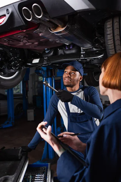 African american mechanic holding wrench while pointing at car near colleague with clipboard in garage — Fotografia de Stock