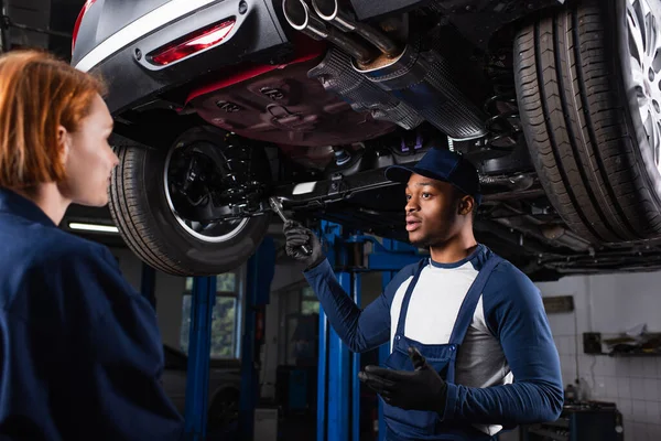 Mecánico afroamericano sosteniendo llave mientras habla con colega debajo del coche en el garaje — Stock Photo