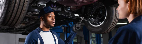 Afroamerikanischer Mechaniker hält Schraubenschlüssel in der Hand, während er mit einem Kollegen im Autoservice spricht, Banner — Stockfoto