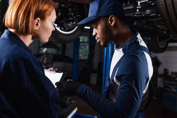 Mecánico afroamericano apuntando a portapapeles cerca de colega en servicio de automóviles - foto de stock