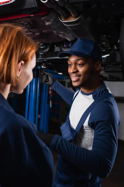 Fröhlicher afrikanisch-amerikanischer Mechaniker im Gespräch mit verschwommenem Kollegen in Uniform im Autoservice — Stockfoto