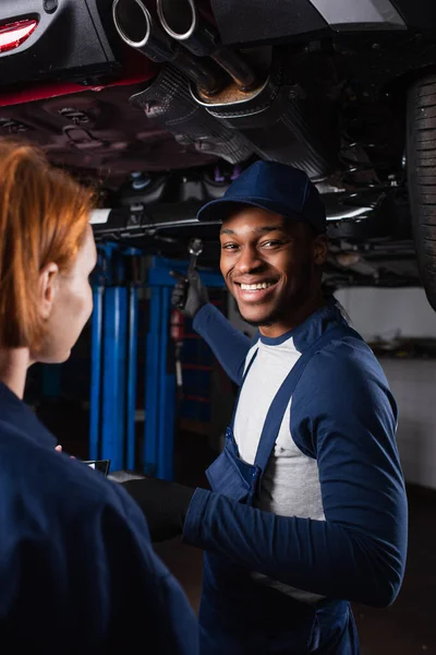 Joyeux ouvrier afro-américain regardant la caméra près de son collègue dans le service de voiture — Photo de stock